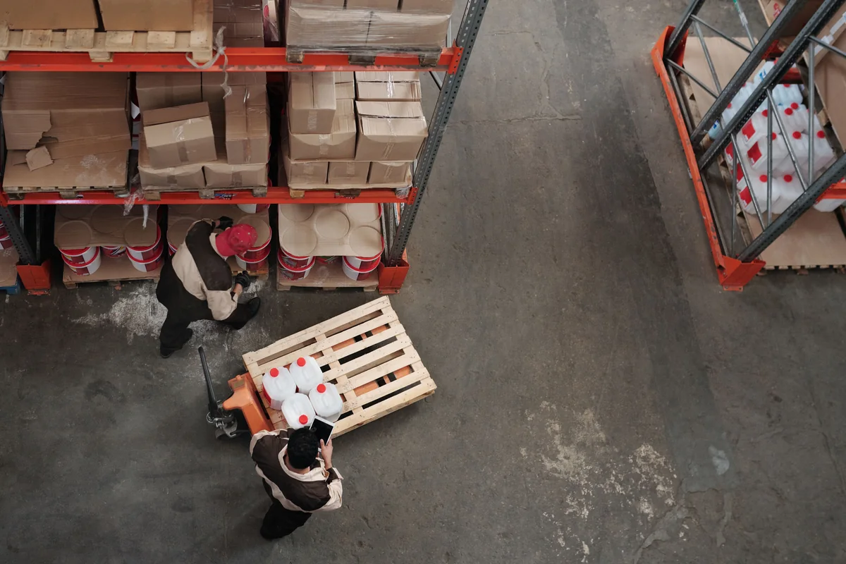 Warehouse workers coordinating inventory among shelves and stacked cartons.